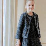A young girl stands indoors by a window, wearing the KIKI and JOJO Girls Vintage Beaded Tweed Jacket in gray—a sophisticated, vintage-inspired look perfect for elegant children's outerwear.