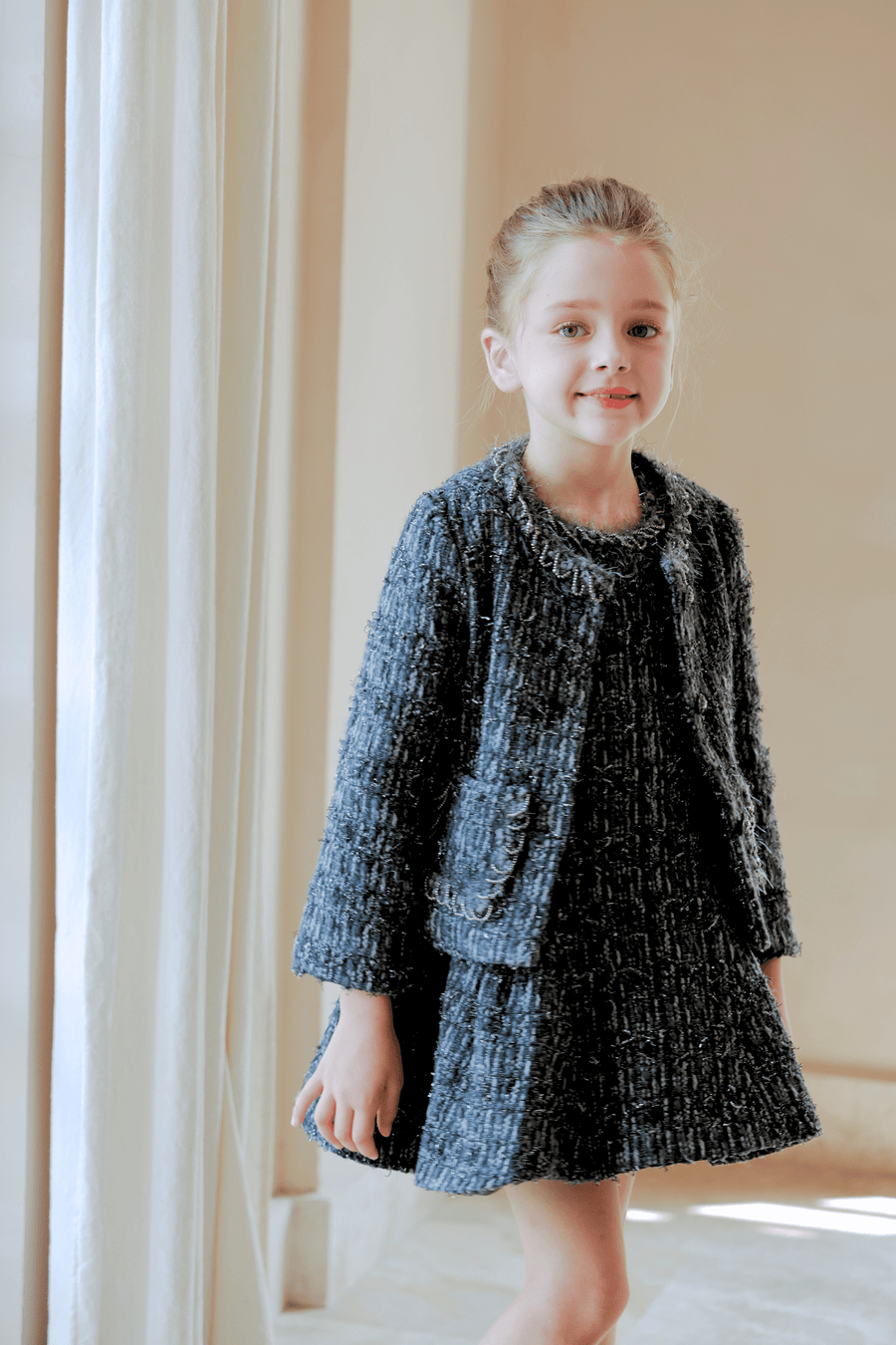 A young girl stands indoors by a window, wearing the KIKI and JOJO Girls Vintage Beaded Tweed Jacket in gray—a sophisticated, vintage-inspired look perfect for elegant children's outerwear.