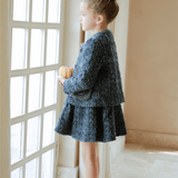 A young girl stands by a window in the KIKI and JOJO Girls Vintage Beaded Tweed Jacket - Gray, holding a small pumpkin and gazing thoughtfully outside.