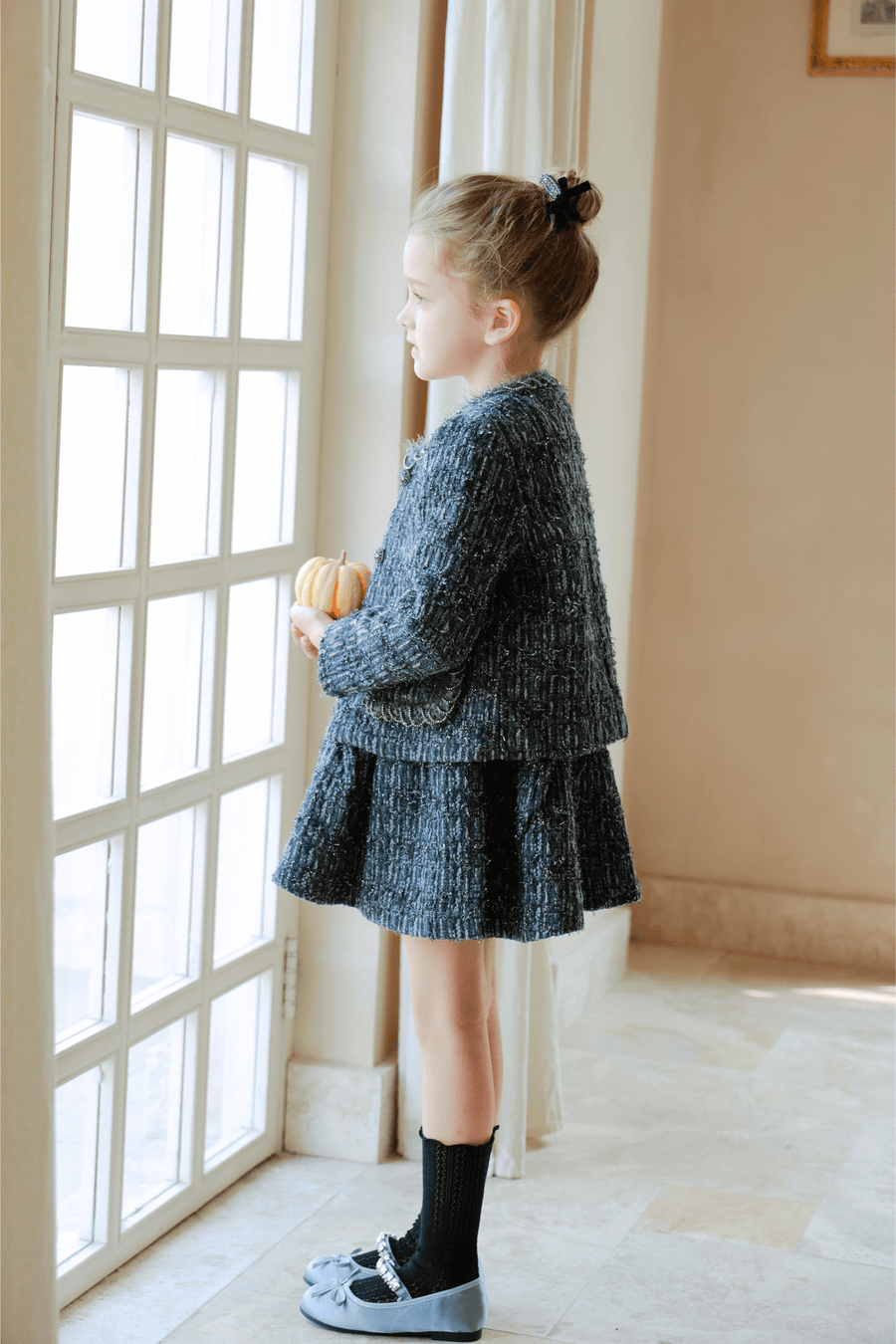 A young girl stands by a window in the KIKI and JOJO Girls Vintage Beaded Tweed Jacket - Gray, holding a small pumpkin and gazing thoughtfully outside.