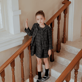 A young girl in the Girls Vintage Beaded Tweed Jacket - Gray by KIKI and JOJO stands on a staircase, hand on railing, looking at the camera and showcasing timeless elegance in sophisticated children's outerwear.
