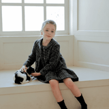 A young girl wearing the KIKI and JOJO Girls Vintage Beaded Tweed Wave-Hem Dress in gray sits on a window bench, petting a small black and white cat.