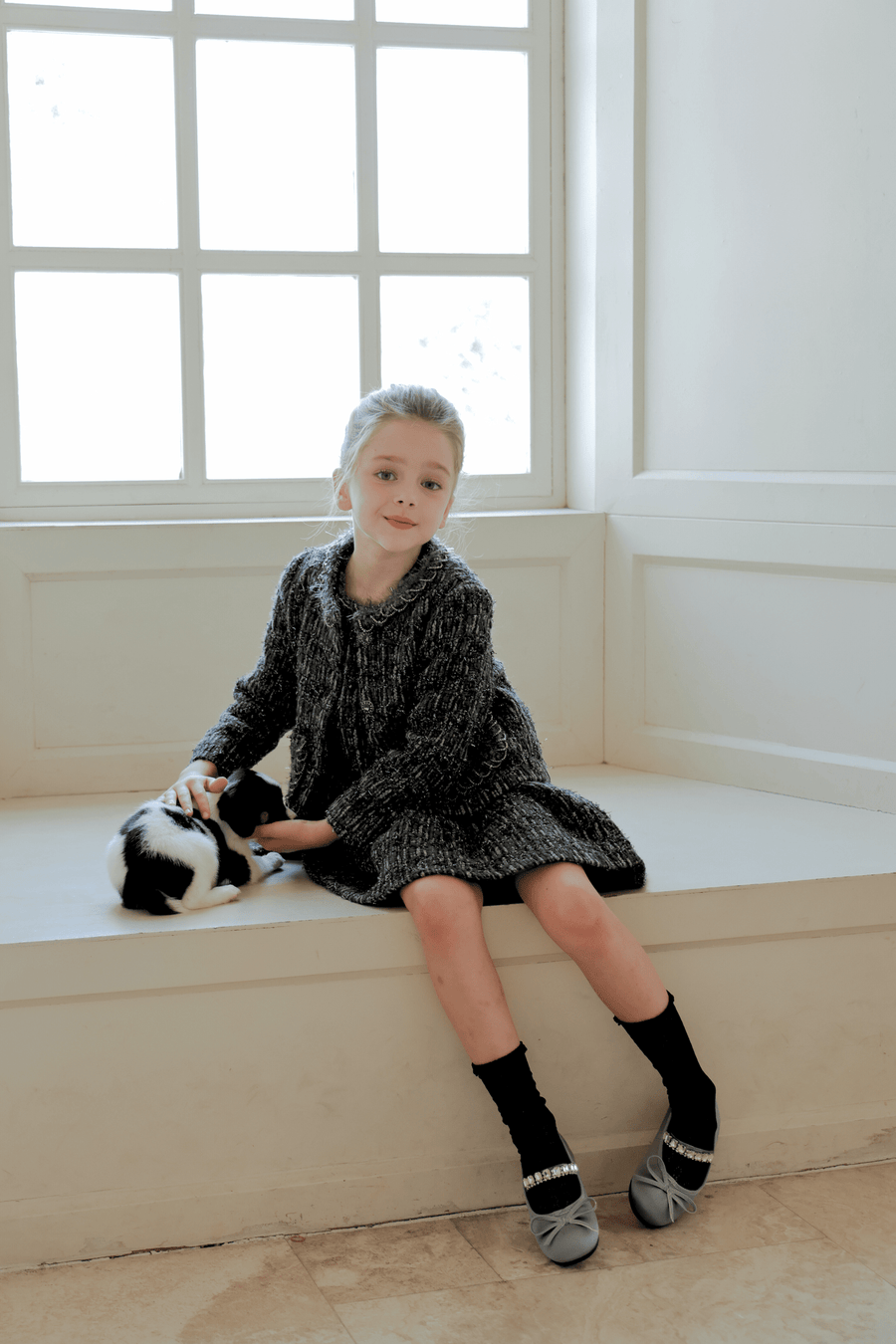 A young girl wearing the KIKI and JOJO Girls Vintage Beaded Tweed Wave-Hem Dress in gray sits on a window bench, petting a small black and white cat.