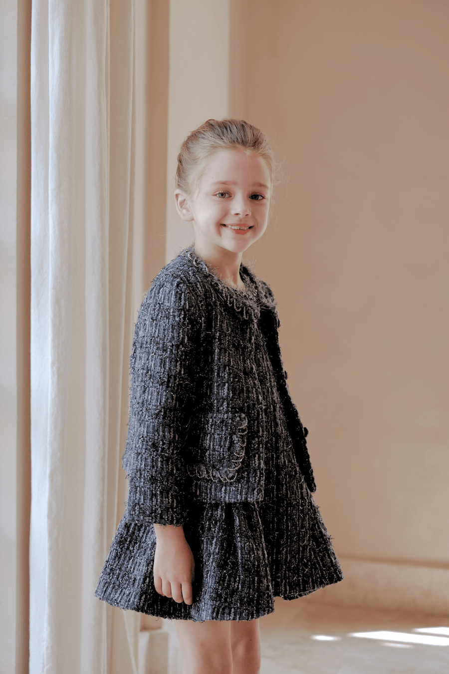 A young girl with light hair smiles at the camera indoors, wearing the KIKI and JOJO Girls Vintage Beaded Tweed Wave-Hem Dress in gray, perfect for special occasions. She stands near a light curtain and beige wall.