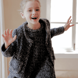A young girl, smiling and raising her hands, wears the KIKI and JOJO Girls Vintage Beaded Tweed Wave-Hem Dress in gray, standing indoors by a window lit with natural light.