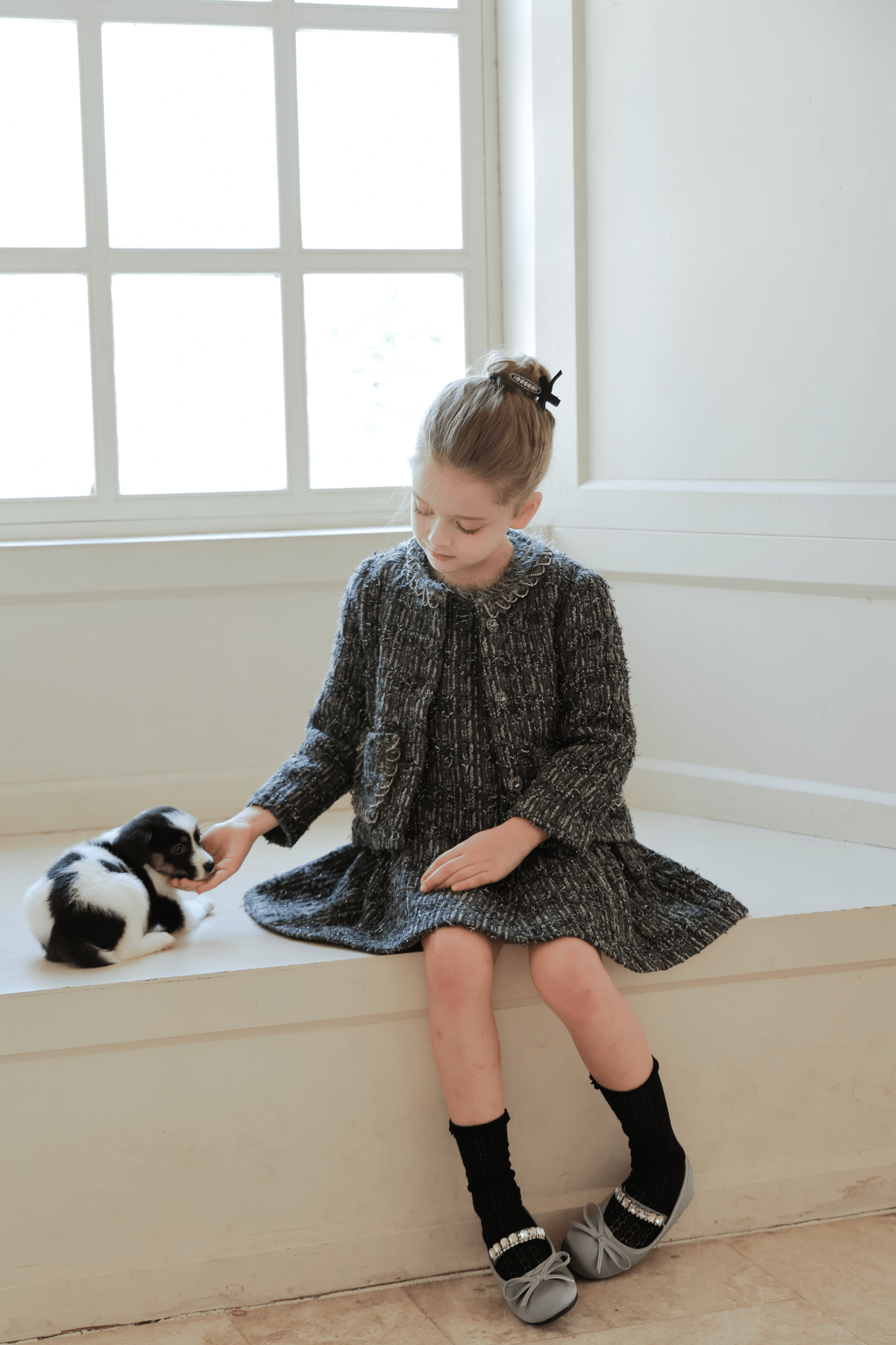 A young girl wears the KIKI and JOJO Girls Vintage Beaded Tweed Wave-Hem Dress in gray as she sits on a windowsill, gently reaching out to a small black and white kitten beside her.