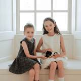 Two young girls sit on a window seat, smiling at the camera. One wears the KIKI and JOJO Girls Vintage Beaded Tweed Wave-Hem Dress in gray and holds a small black and white kitten on her lap.