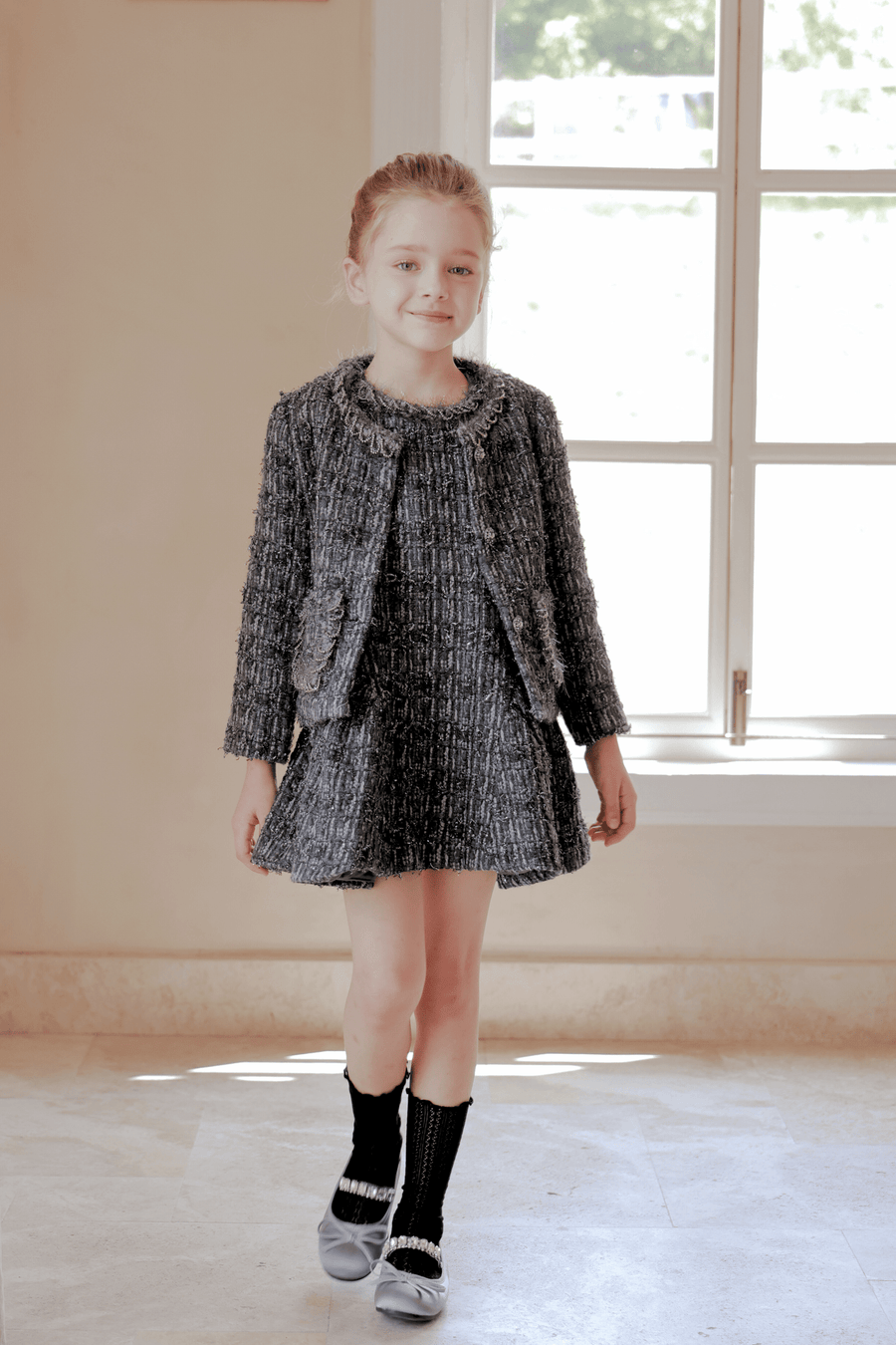 A young girl stands indoors in front of a window, wearing the KIKI and JOJO Girls Vintage Beaded Tweed Wave-Hem Dress in gray—a chic choice for special occasions.