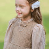 A young girl with long brown hair wears the KIKI and JOJO Girls Vintage Elegance Embroidered Velvet & Tulle Dress and a white hair accessory, standing outdoors on a sunny day.