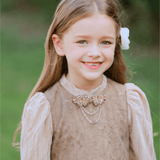A young girl wearing the KIKI and JOJO Girls Vintage Elegance Embroidered Velvet & Tulle Dress stands outdoors, smiling with a white flower in her hair, surrounded by greenery.
