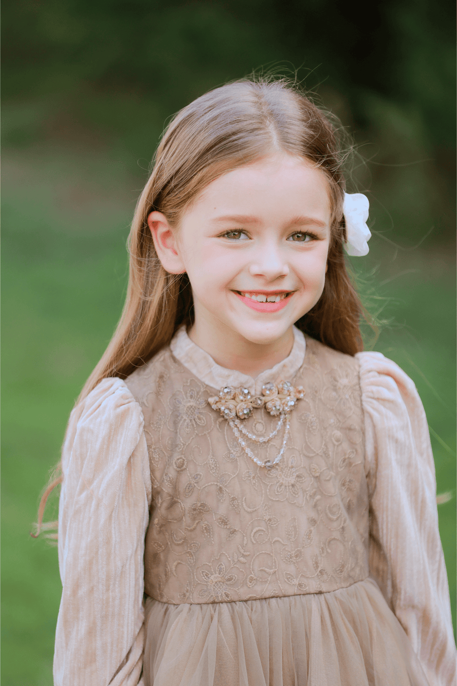 A young girl wearing the KIKI and JOJO Girls Vintage Elegance Embroidered Velvet & Tulle Dress stands outdoors, smiling with a white flower in her hair, surrounded by greenery.