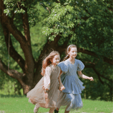 Two young girls wear KIKI and JOJO’s Girls Vintage Elegance Embroidered Velvet & Tulle Dress as they run hand in hand on grass, with trees creating a picturesque backdrop.