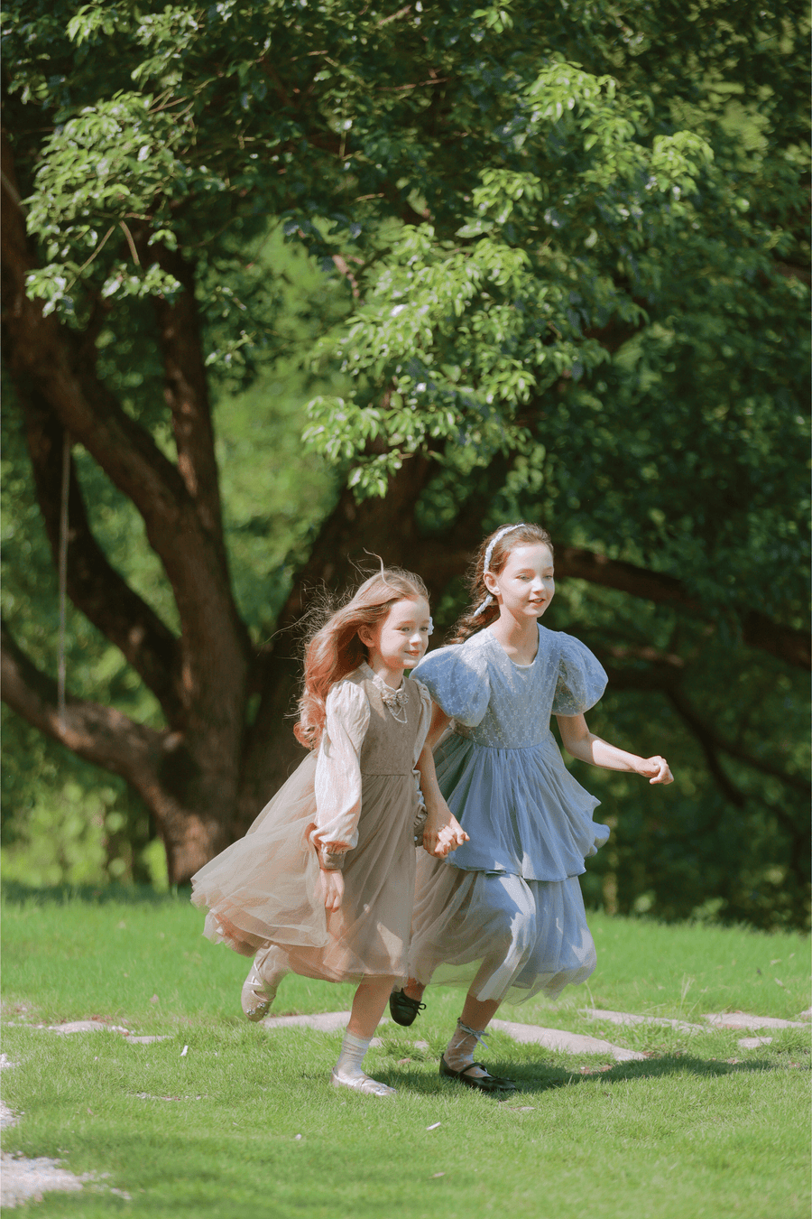 Two young girls wear KIKI and JOJO’s Girls Vintage Elegance Embroidered Velvet & Tulle Dress as they run hand in hand on grass, with trees creating a picturesque backdrop.