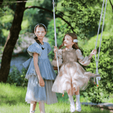 Two girls in KIKI and JOJO's Vintage Elegance Embroidered Velvet & Tulle Dress enjoy a sunny day outdoors on a swing, surrounded by green grass and trees—an ideal choice for formal occasions.