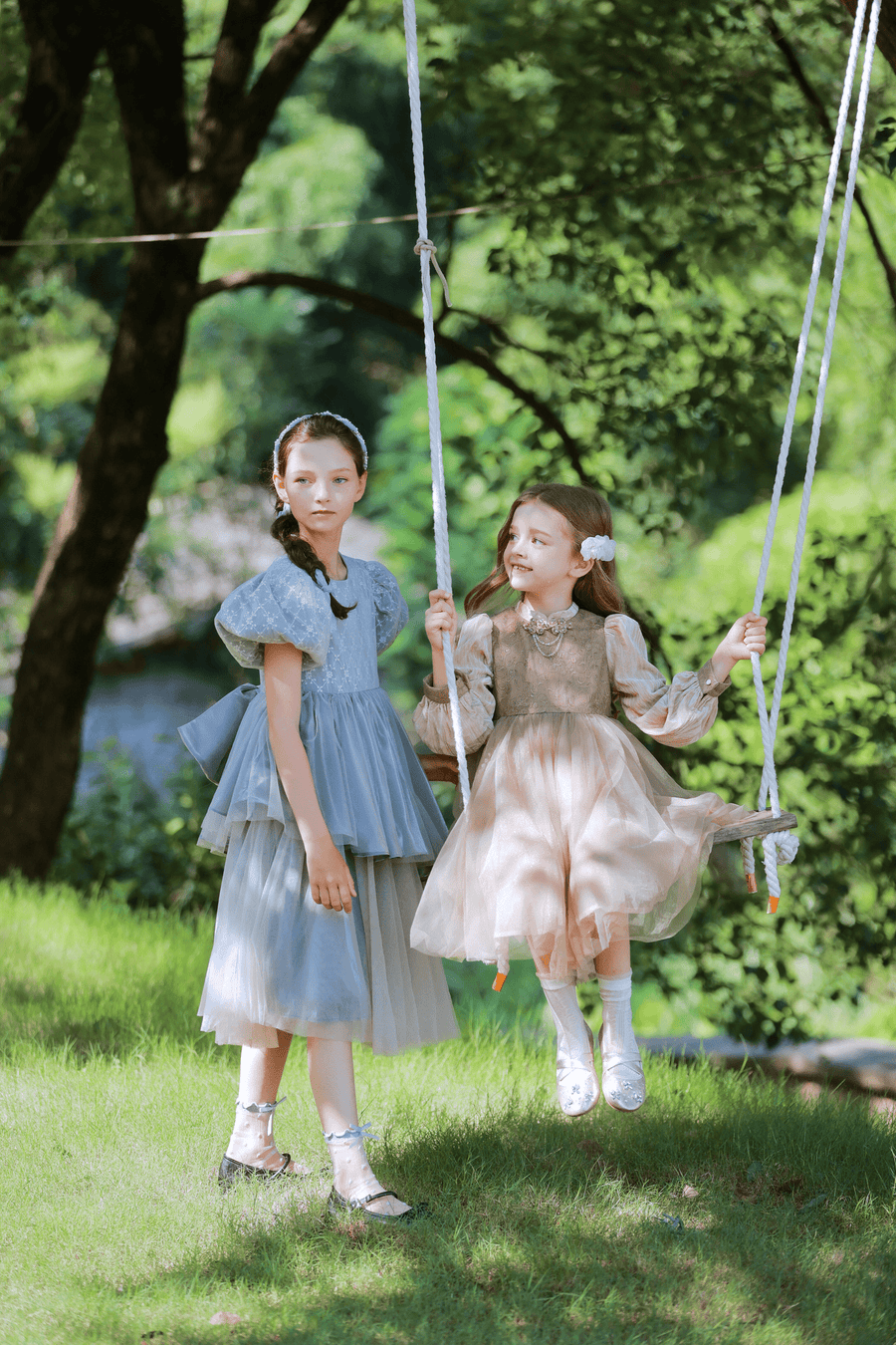 Two girls in KIKI and JOJO's Vintage Elegance Embroidered Velvet & Tulle Dress enjoy a sunny day outdoors on a swing, surrounded by green grass and trees—an ideal choice for formal occasions.