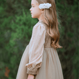 A young girl in profile wears the KIKI and JOJO Girls Vintage Elegance Embroidered Velvet & Tulle Dress, featuring floral embroidery and a white hair accessory, standing outdoors against a green, leafy background—ideal for formal occasions.