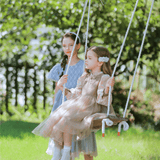 Two young girls wear KIKI and JOJO’s Girls Vintage Elegance Embroidered Velvet & Tulle Dress in a garden—one sits on a wooden swing while the other stands behind, both showcasing the dress's beautiful floral embroidery.