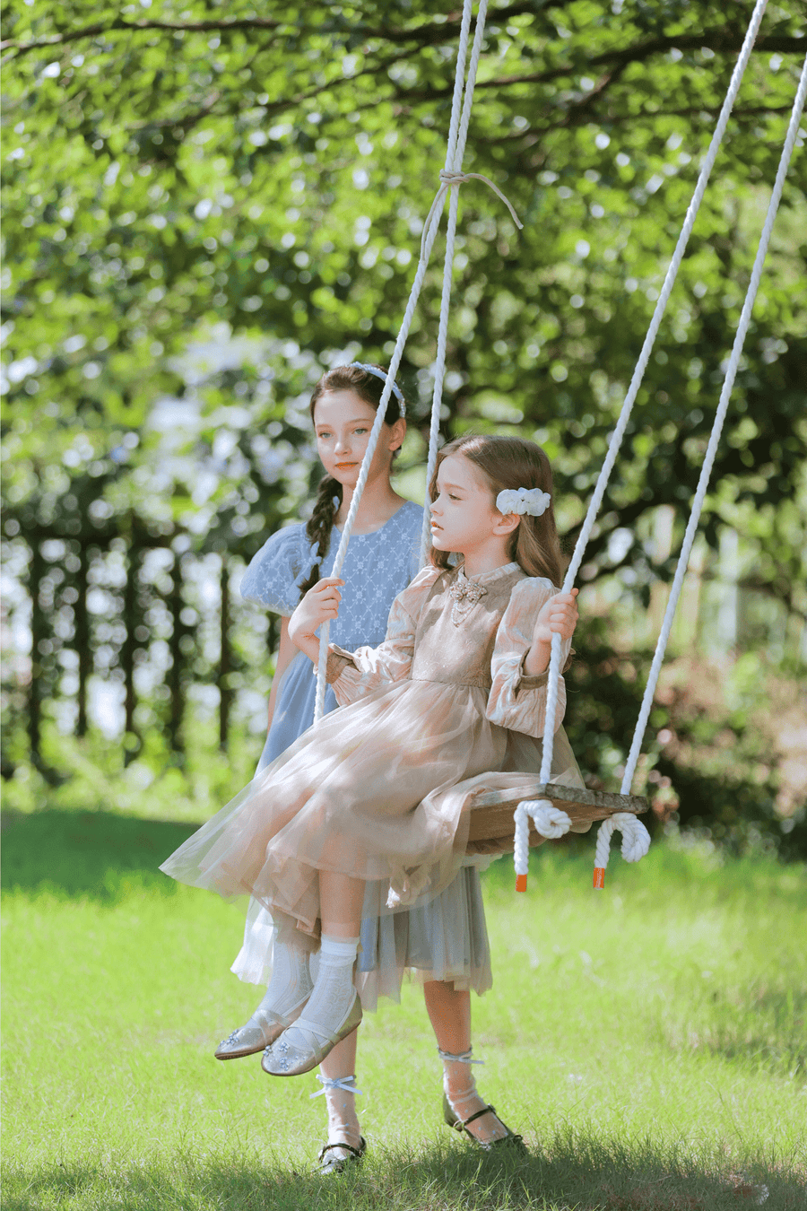Two young girls wear KIKI and JOJO’s Girls Vintage Elegance Embroidered Velvet & Tulle Dress in a garden—one sits on a wooden swing while the other stands behind, both showcasing the dress's beautiful floral embroidery.