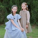 Two girls pose outdoors in KIKI and JOJO’s Girls Vintage Elegance Embroidered Velvet & Tulle Dresses—one seated in light blue, the other standing in beige. Both have long hair and neutral expressions.