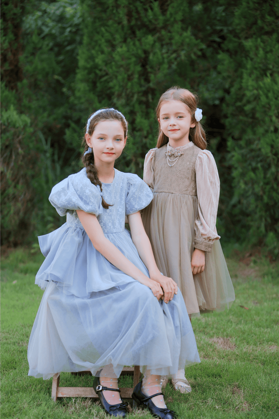 Two girls pose outdoors in KIKI and JOJO’s Girls Vintage Elegance Embroidered Velvet & Tulle Dresses—one seated in light blue, the other standing in beige. Both have long hair and neutral expressions.