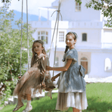 Two young girls wear KIKI and JOJO’s Girls Vintage Elegance Embroidered Velvet & Tulle Dress on a wooden swing in a sunny garden, with a large white house in the background.