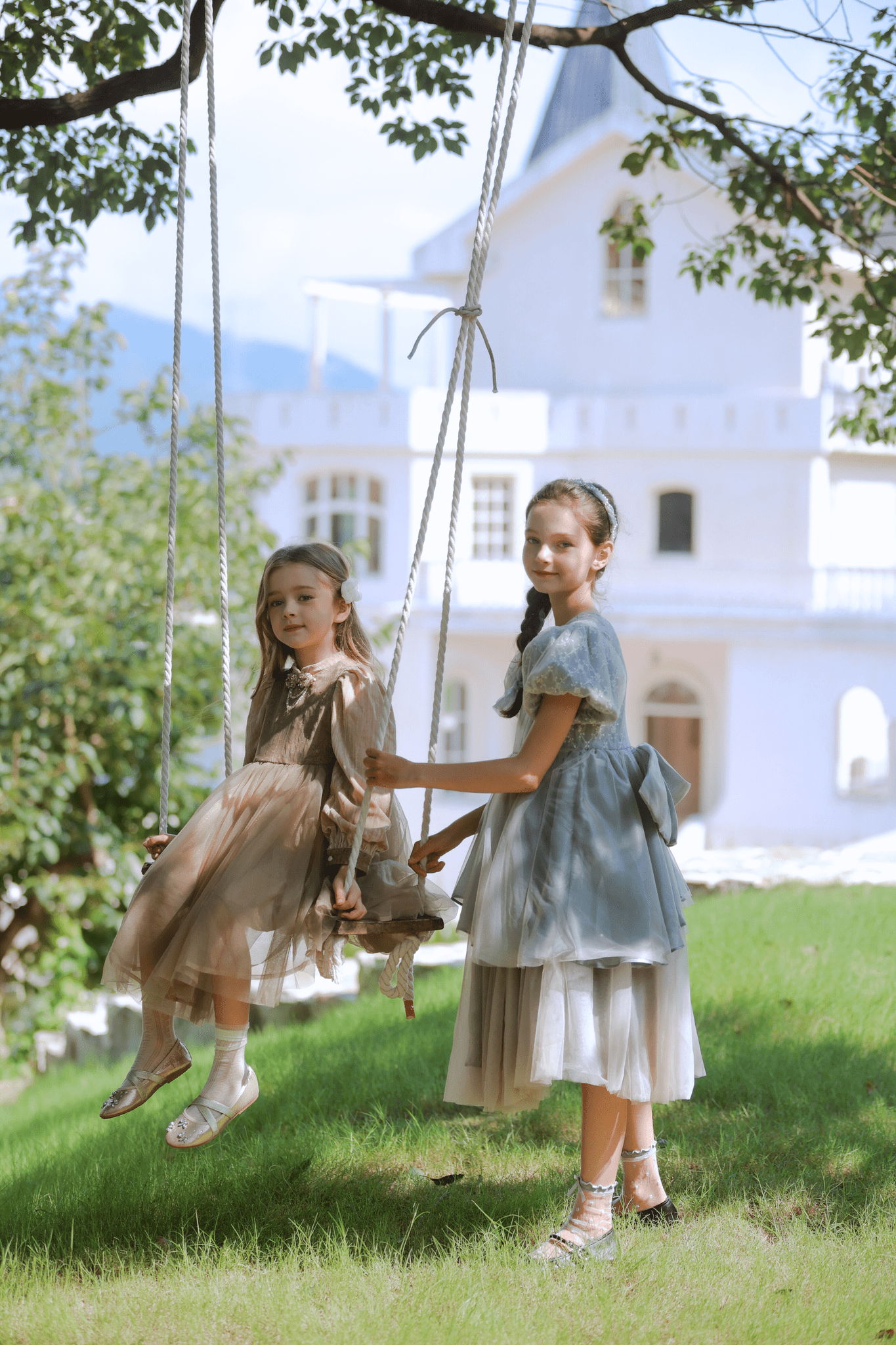 Two young girls wear KIKI and JOJO’s Girls Vintage Elegance Embroidered Velvet & Tulle Dress on a wooden swing in a sunny garden, with a large white house in the background.