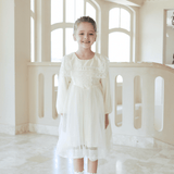 A young girl smiles by large windows wearing the KIKI and JOJO Girls Vintage Lace Satin & Tulle Dress in beige, styled with white socks and shoes.