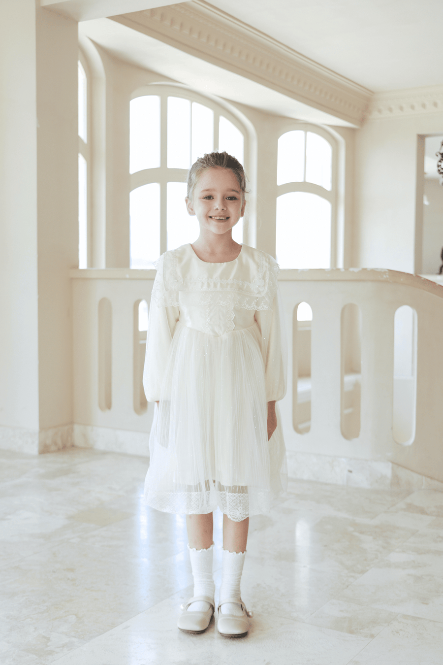 A young girl smiles by large windows wearing the KIKI and JOJO Girls Vintage Lace Satin & Tulle Dress in beige, styled with white socks and shoes.