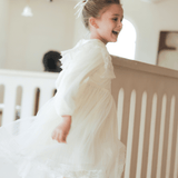 A young girl in the KIKI and JOJO Girls Vintage Lace Satin & Tulle Dress in beige, featuring hand-embroidered floral details, runs indoors with a smile, her hair tied back with a white ribbon near a light-colored railing.