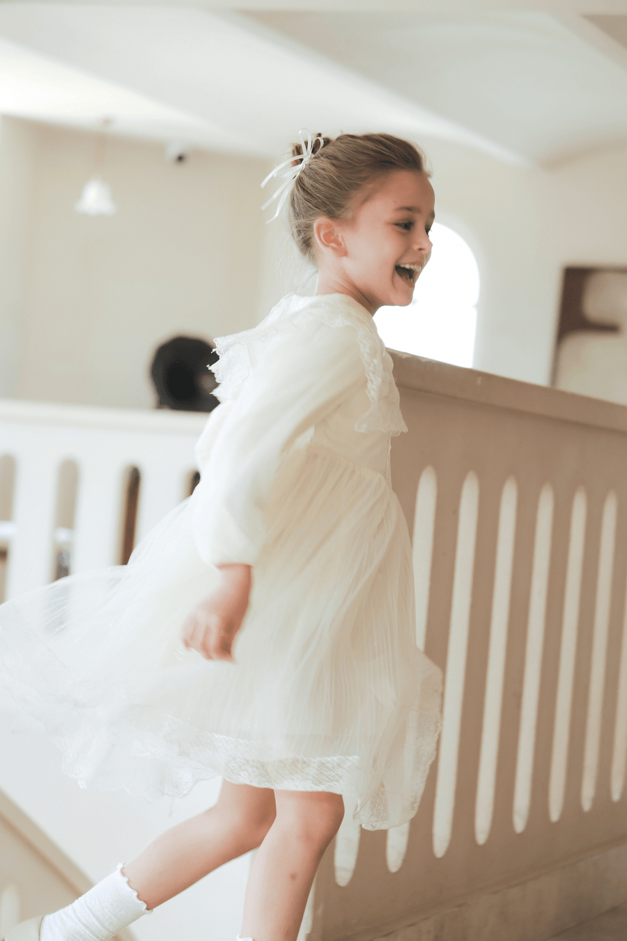 A young girl in the KIKI and JOJO Girls Vintage Lace Satin & Tulle Dress in beige, featuring hand-embroidered floral details, runs indoors with a smile, her hair tied back with a white ribbon near a light-colored railing.