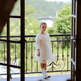A young girl wears the KIKI and JOJO Girls Vintage Lace Satin & Tulle Dress in beige, standing on a balcony with black railings, overlooking green trees and a river through open double doors.