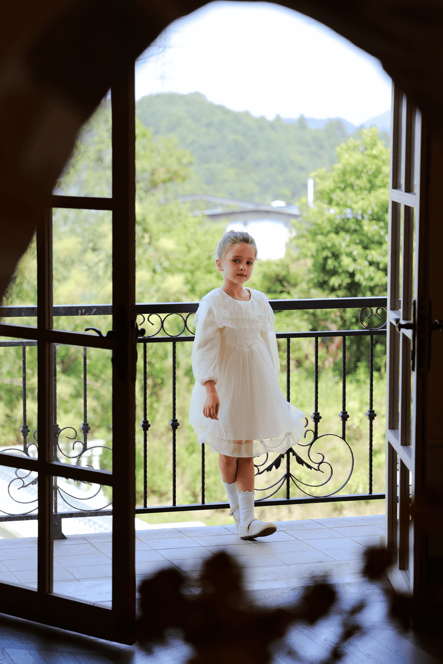 A young girl wears the KIKI and JOJO Girls Vintage Lace Satin & Tulle Dress in beige, standing on a balcony with black railings, overlooking green trees and a river through open double doors.