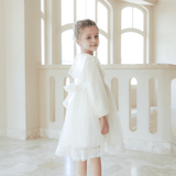 A young girl wears the KIKI and JOJO Girls Vintage Lace Satin & Tulle Dress in beige, standing indoors with a slight smile as light from large windows highlights the delicate bows on her socks.