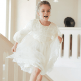 A young girl, smiling, runs down an indoor staircase in the KIKI and JOJO Girls Vintage Lace Satin & Tulle Dress - Beige, with soft natural light streaming through large windows.