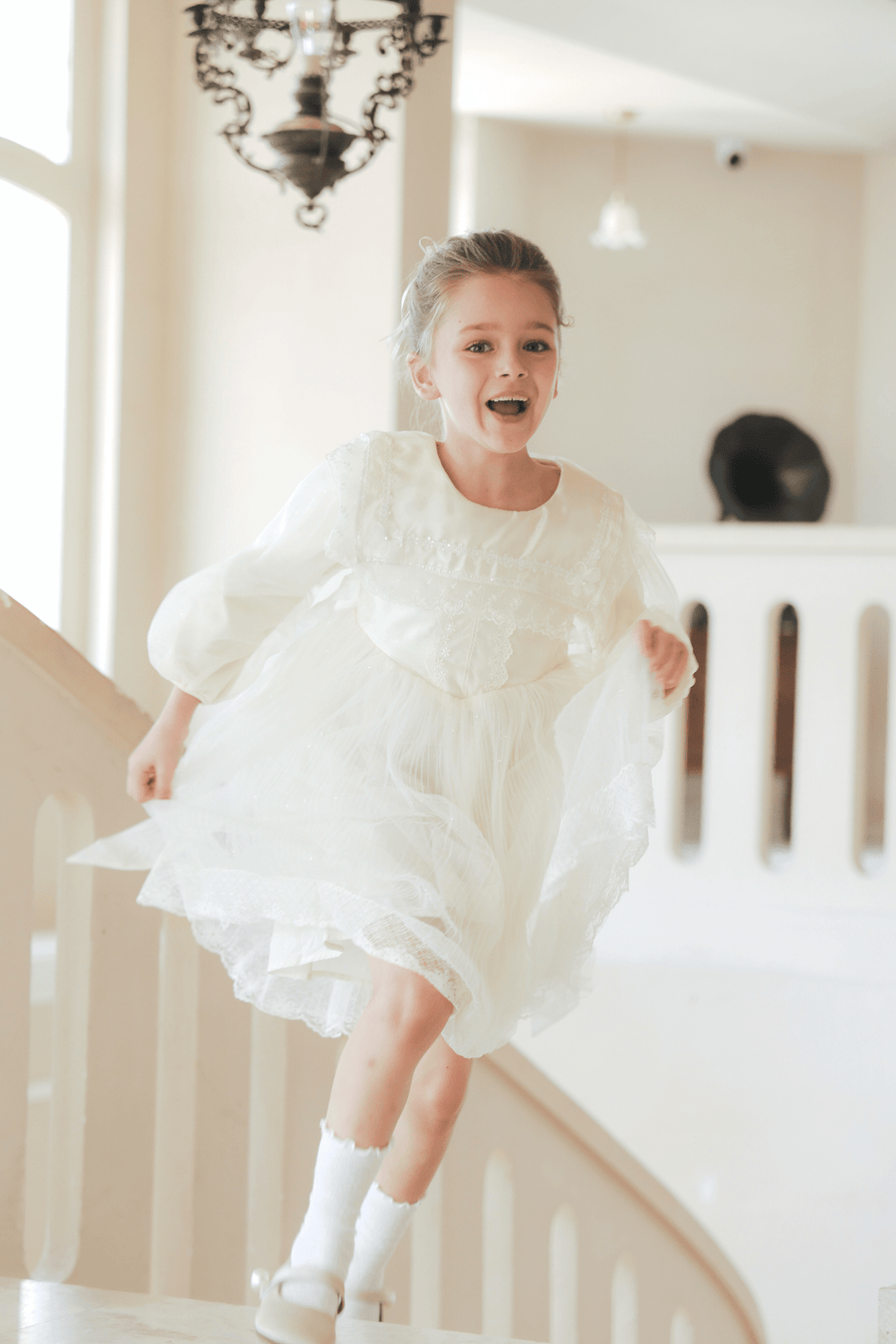 A young girl, smiling, runs down an indoor staircase in the KIKI and JOJO Girls Vintage Lace Satin & Tulle Dress - Beige, with soft natural light streaming through large windows.