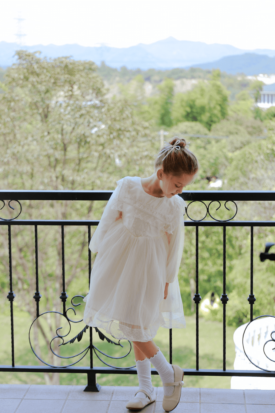 A young girl wearing the KIKI and JOJO Girls Vintage Lace Satin & Tulle Dress in beige stands on a balcony, looking down, with trees and mountains in the background.
