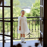 A young girl wears the KIKI and JOJO Girls Vintage Lace Satin & Tulle Dress in beige, featuring hand-embroidered floral details, as she stands on a balcony with greenery and a bridge visible through open glass doors.