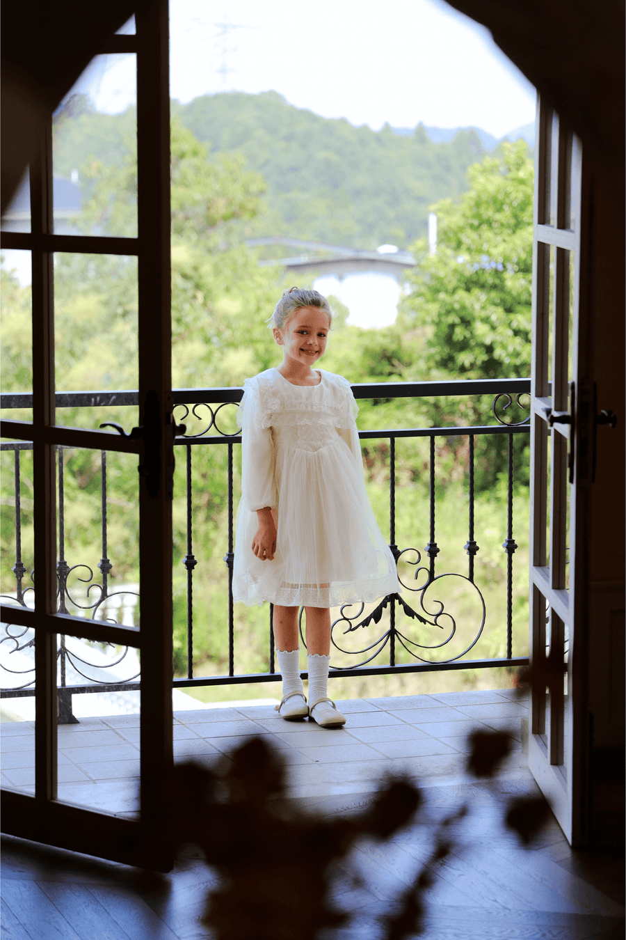 A young girl wears the KIKI and JOJO Girls Vintage Lace Satin & Tulle Dress in beige, featuring hand-embroidered floral details, as she stands on a balcony with greenery and a bridge visible through open glass doors.
