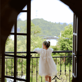 A young girl in the KIKI and JOJO Girls Vintage Lace Satin & Tulle Dress in beige stands on a balcony, gazing at trees and distant hills through an open arched doorway.