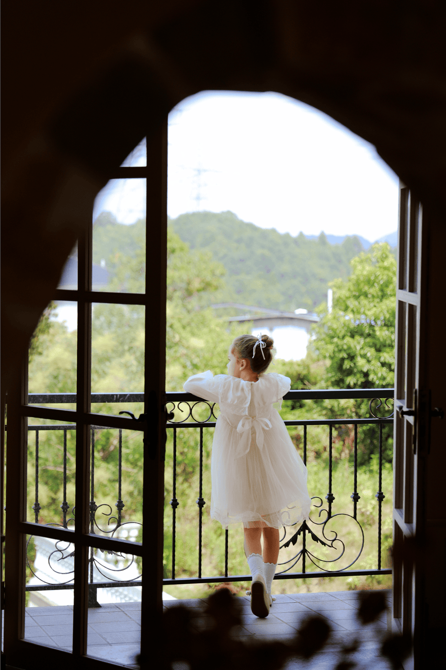 A young girl in the KIKI and JOJO Girls Vintage Lace Satin & Tulle Dress in beige stands on a balcony, gazing at trees and distant hills through an open arched doorway.