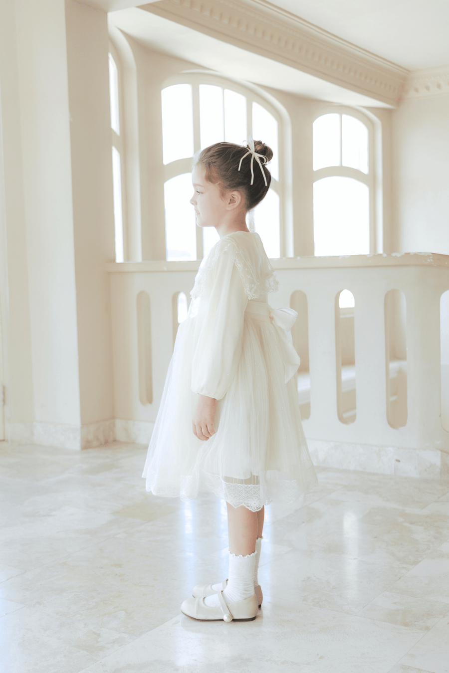 A girl stands indoors in profile, wearing the KIKI and JOJO Girls Vintage Lace Satin & Tulle Dress in beige, with natural light filtering through large windows behind her.