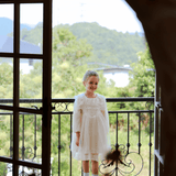 A girl in the KIKI and JOJO Girls Vintage Lace Satin & Tulle Dress - Beige stands on a balcony by an open doorway, with trees and hills in the background.