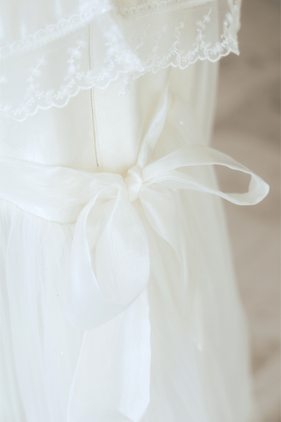 Close-up of the KIKI and JOJO Girls Vintage Lace Satin & Tulle Dress in beige, featuring white lace details, satin tulle, and a sheer fabric bow at the waist.