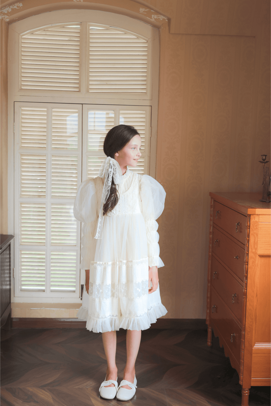 A young girl wears the KIKI and JOJO Girls Vintage Pearl Embroidered Tulle Dress in beige, featuring puffed sleeves and an embroidered bodice, as she stands indoors near a window and wooden dresser, looking to the side.