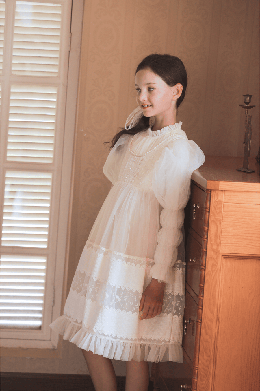 A young girl wears the KIKI and JOJO Girls Vintage Pearl Embroidered Tulle Dress in beige as she stands indoors, leaning against a wooden dresser near a window with shutters.