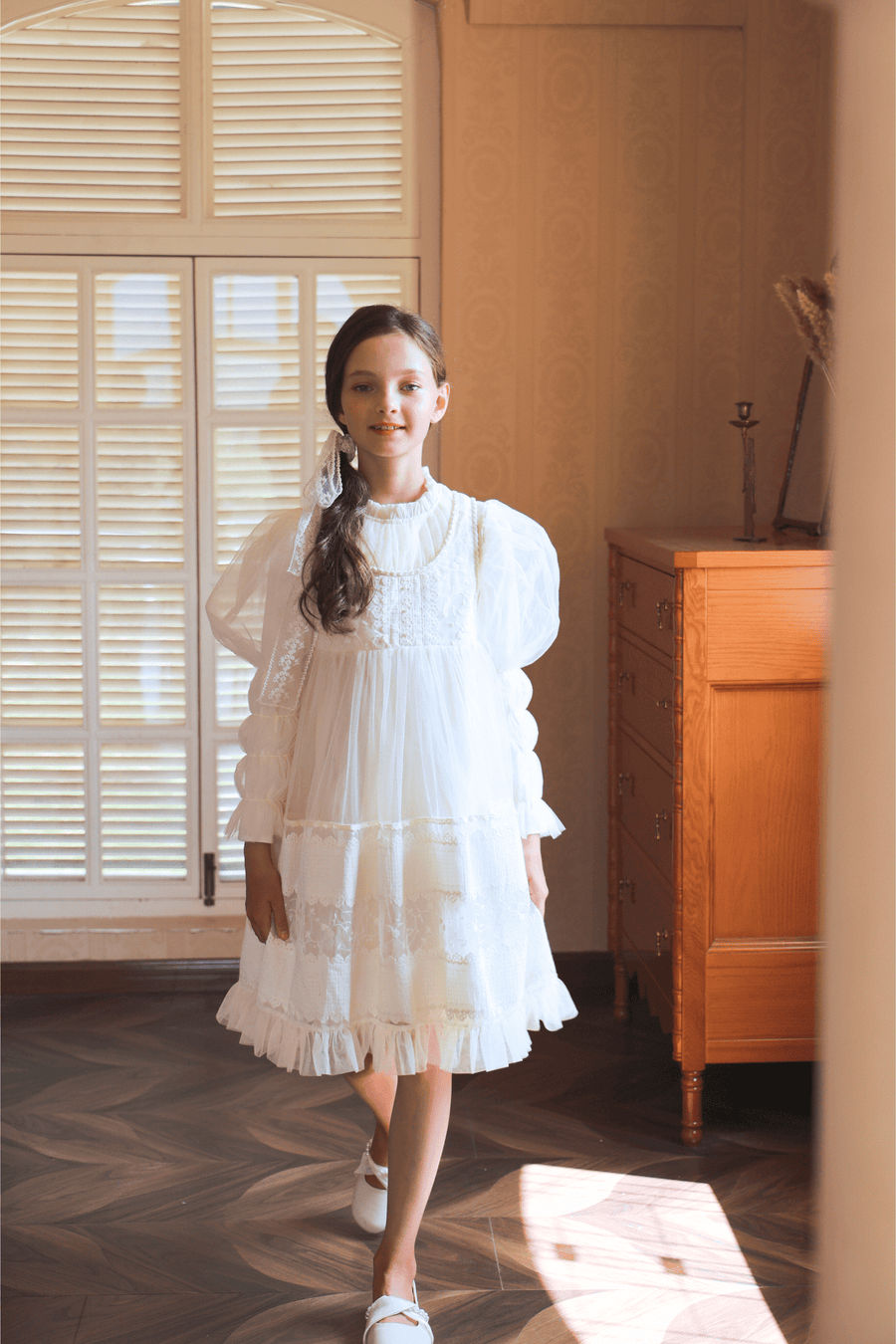 A girl wearing the KIKI and JOJO Girls Vintage Pearl Embroidered Tulle Dress in beige stands indoors on a wooden floor near a window and dresser.