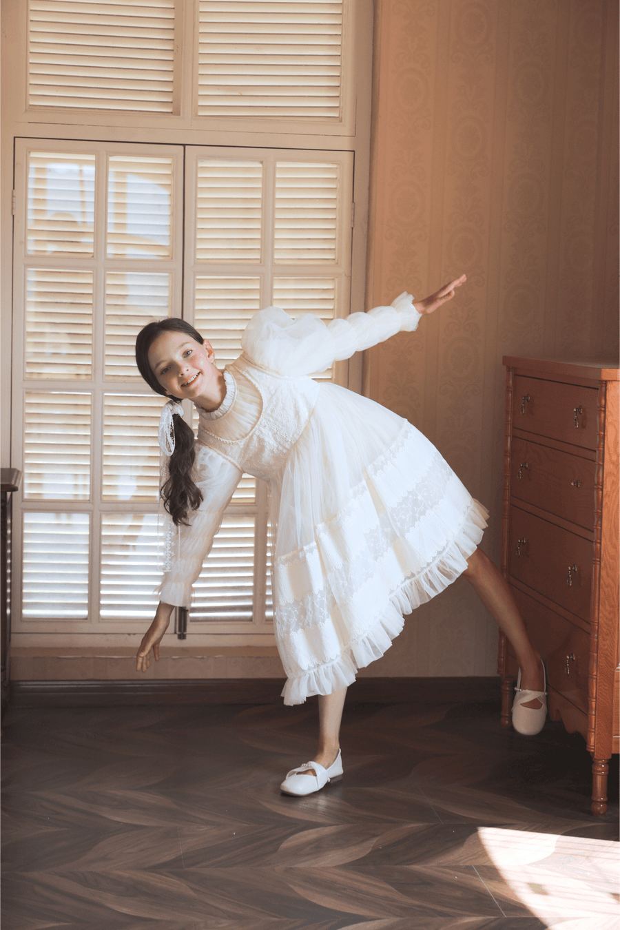 A young girl wears the KIKI and JOJO Girls Vintage Pearl Embroidered Tulle Dress in beige, posing indoors with arms outstretched beside a wooden dresser and a window with closed shutters.