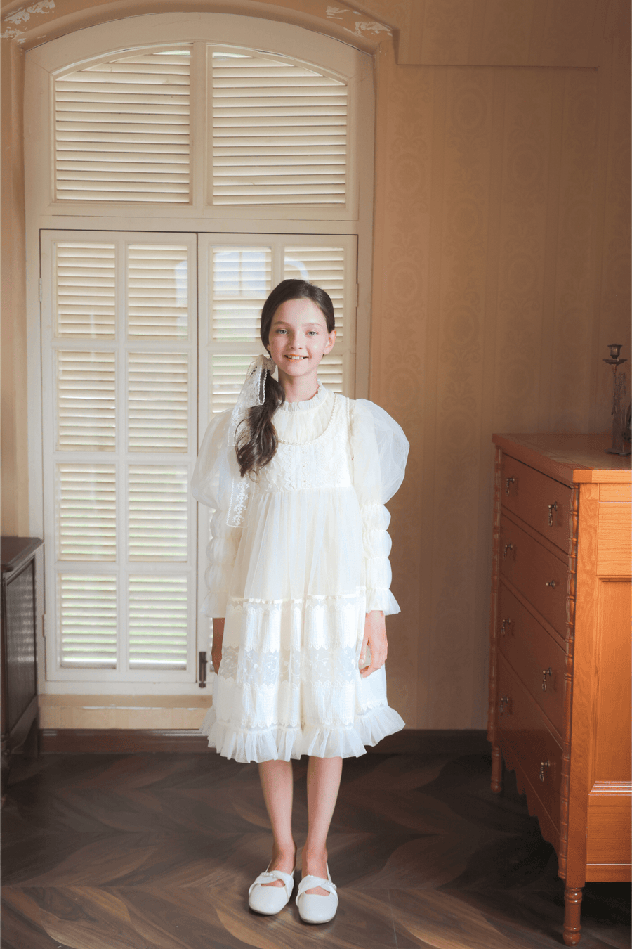 A young girl smiles at the camera indoors, wearing the KIKI and JOJO Girls Vintage Pearl Embroidered Tulle Dress in beige, featuring an embroidered bodice and lace details, paired with white shoes.