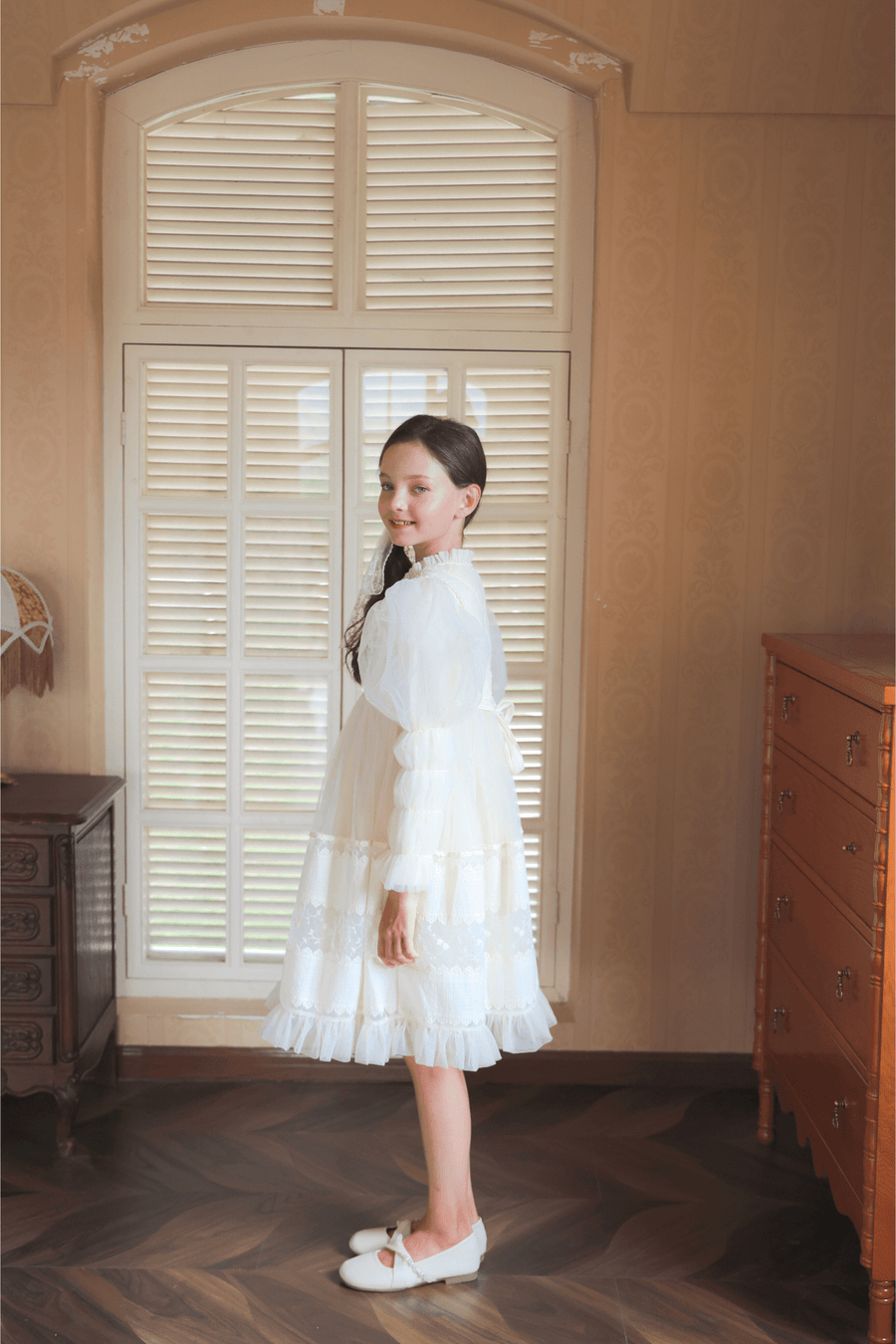A girl wears the KIKI and JOJO Girls Vintage Pearl Embroidered Tulle Dress in beige, standing sideways by a window with closed shutters in a softly lit, vintage-style room.