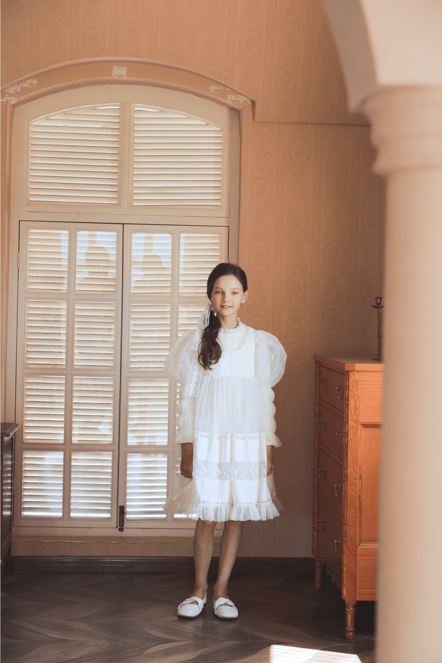 A girl wears the KIKI and JOJO Girls Vintage Pearl Embroidered Tulle Dress in beige, standing indoors by a large window with closed shutters, beside a wooden dresser on a patterned floor.